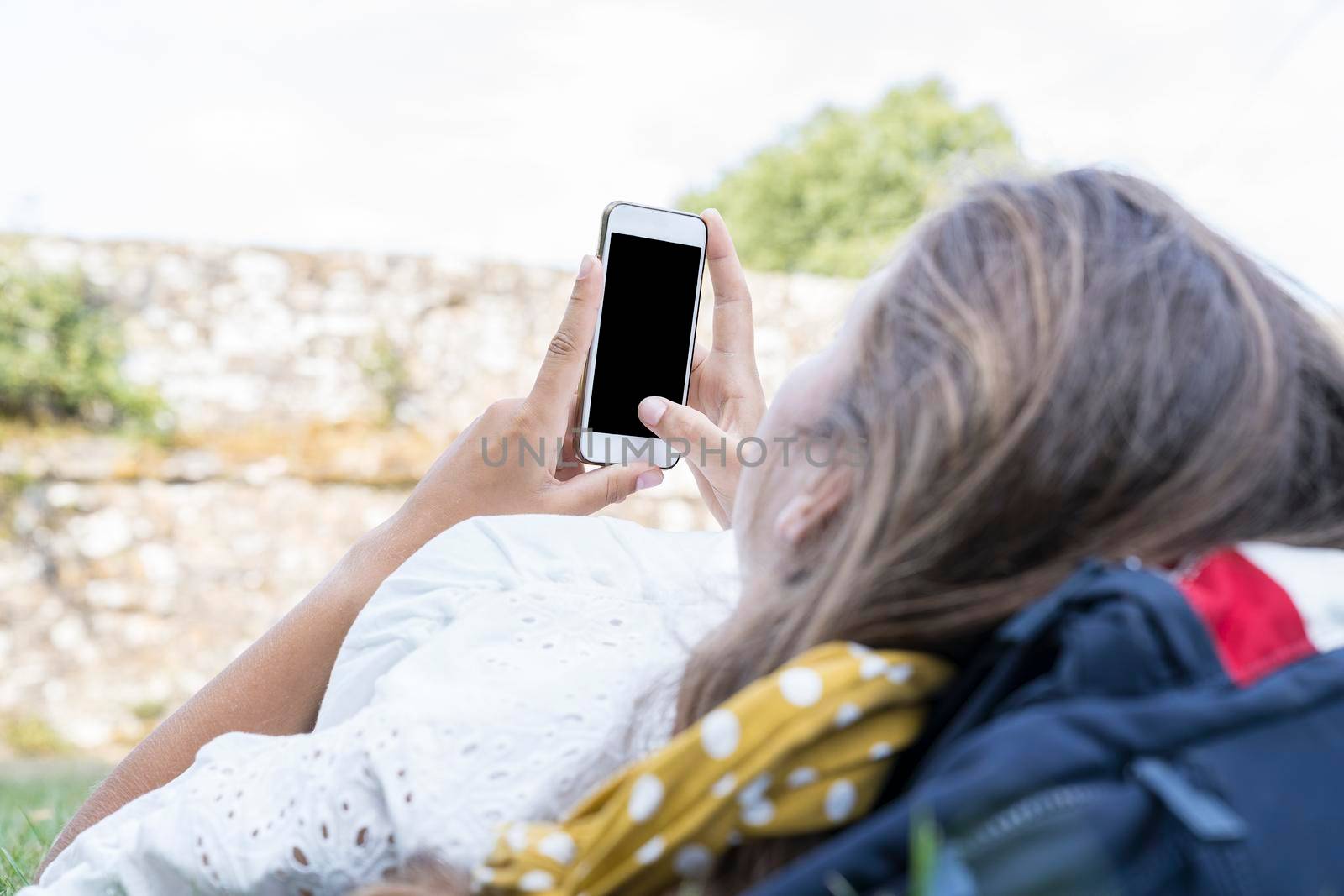 Mockup image of unrecognizable young womans hands holding white mobile phone with blank black screen held up in hands while lying and chilling in the grass with copy space by LeoniekvanderVliet