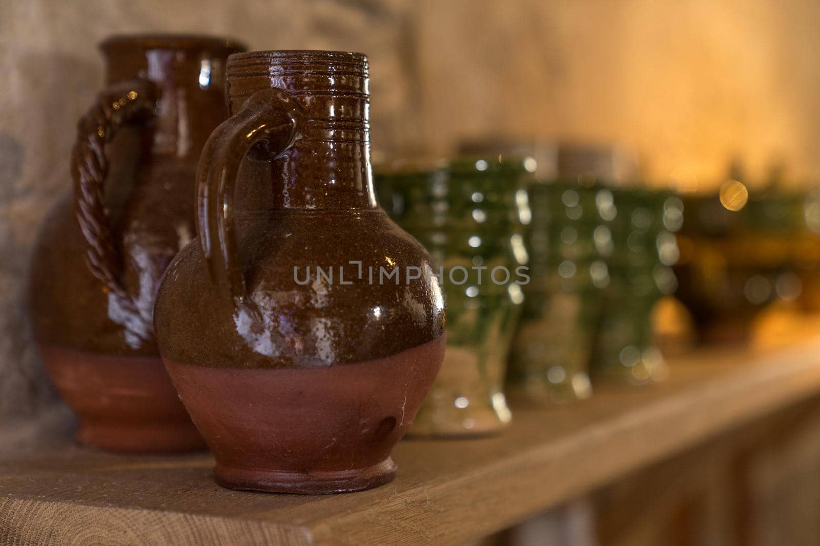 Many medieval earthenware jugs and cups in brown and green glazing standing on a wooden shelf near stone wall