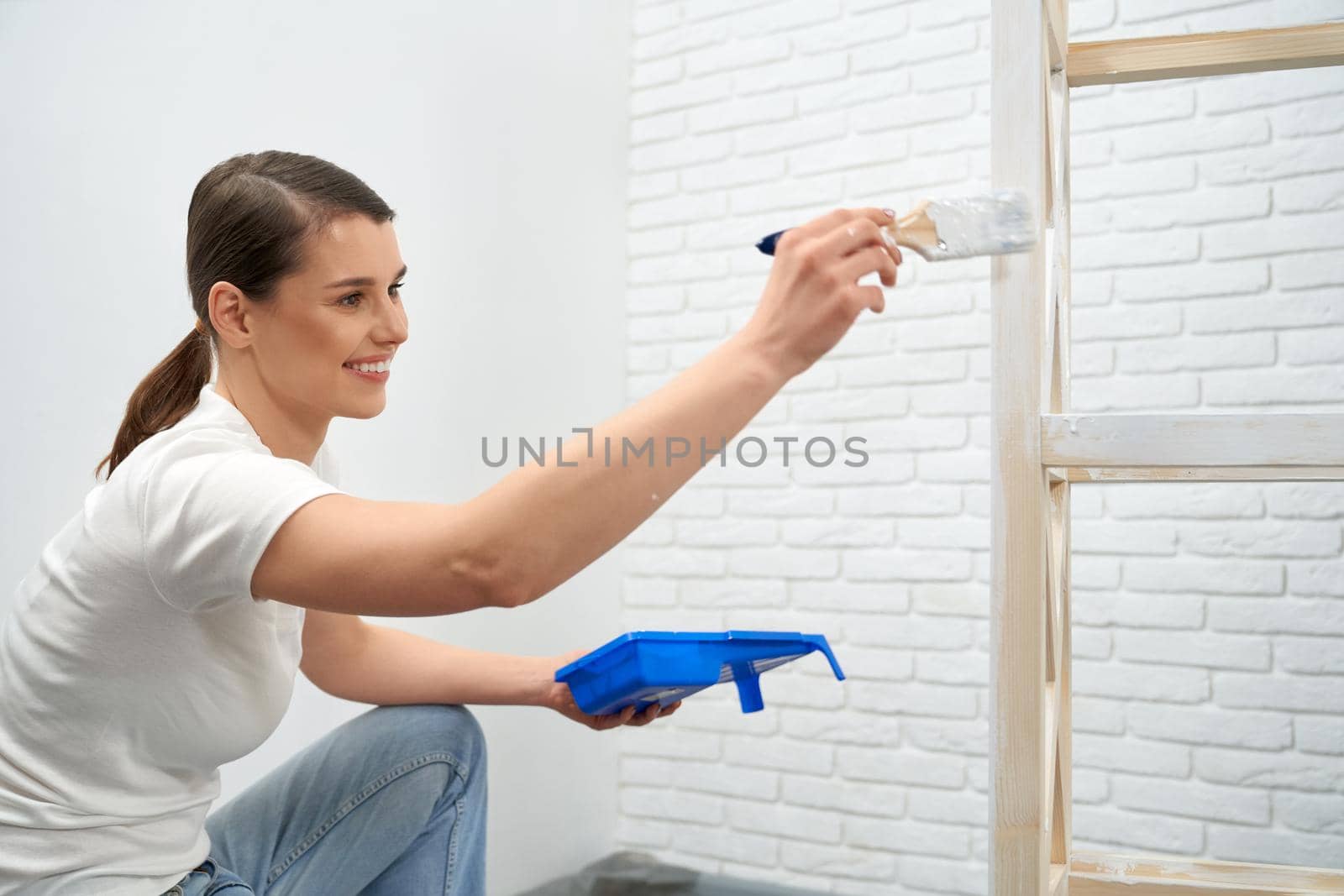 Side view of smiling brunette woman painting wooden rack in white color at home. Concept of improvement wooden rack.