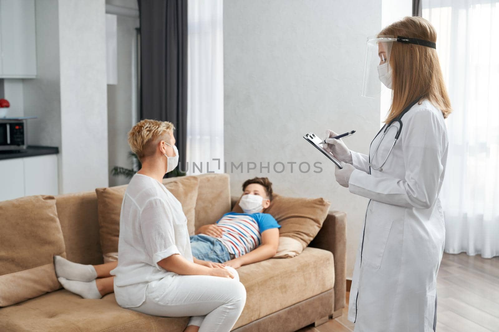 Caring mother sitting on couch near her sick son while competent doctor in medical uniform standing near and writing on clipboard. Pediatrician visiting patient at home.