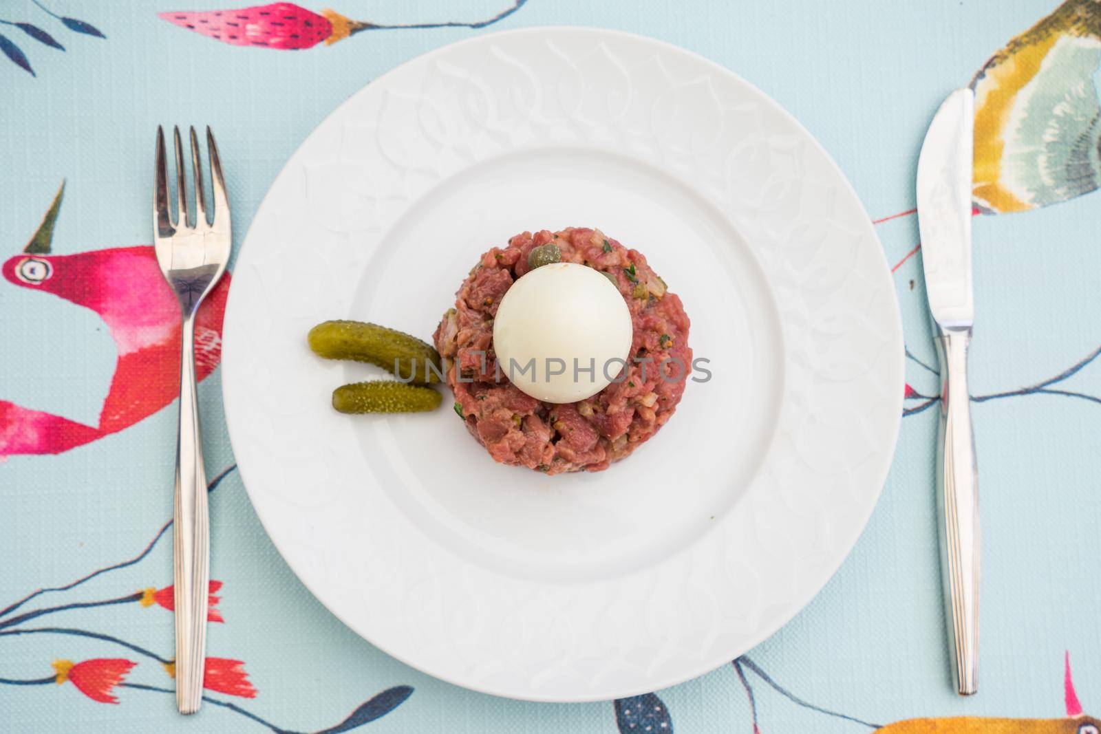 Raw steak tatar with a boiled egg and gurkins on a white plate with cuttlery on a decorative set table