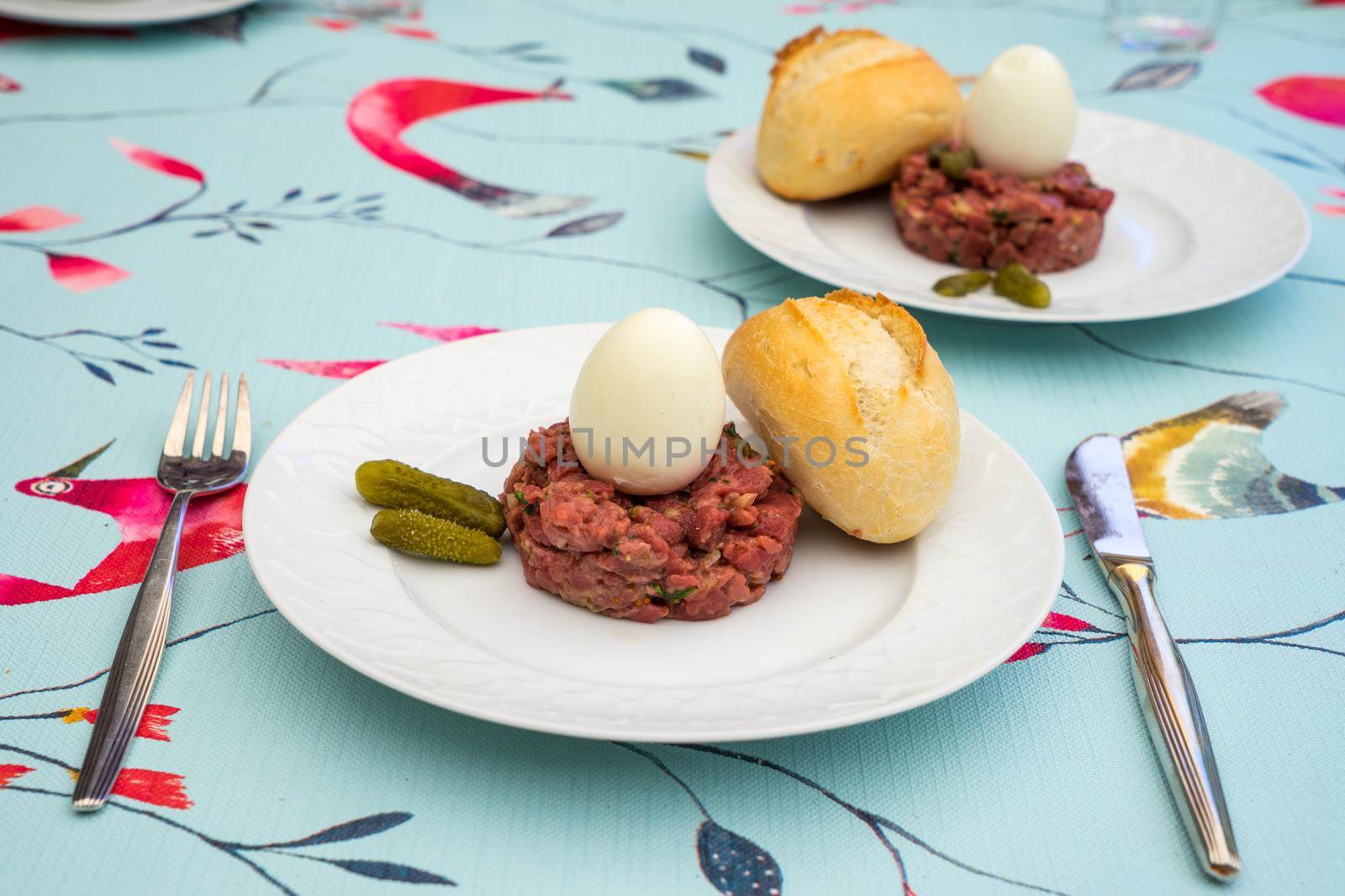 Raw steak tatar with a boiled egg and gurkins on a white plate with cuttlery on a decorative set table