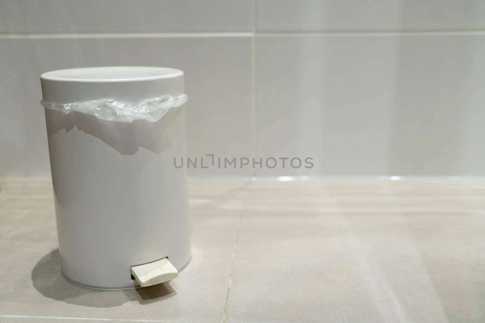 A closed white pedal bin standing on a tiled floor in a bathroom, concept for tidiness and the environment