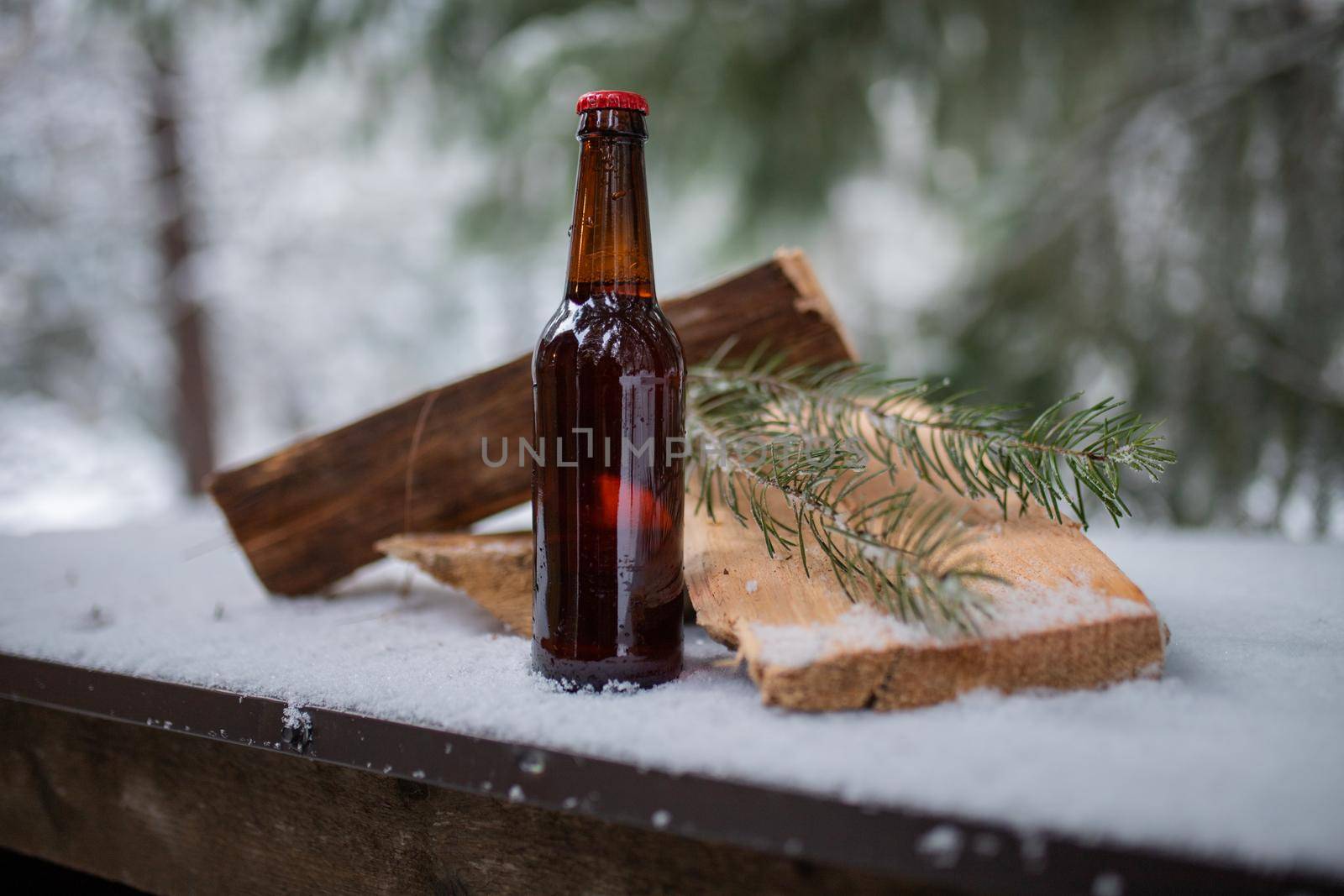 Bottle of beer, wood planks, and pine leaves on a snow-covered table with blurry forest as background. Bottle of alcoholic beverage and snowy pine trees. Winter still life photography