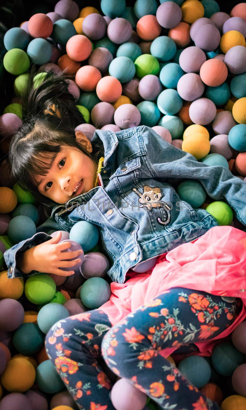 Little asian girl having fun in ball pit with colorful balls. Child playing on indoor playground. Kid jumping in ball pool.
