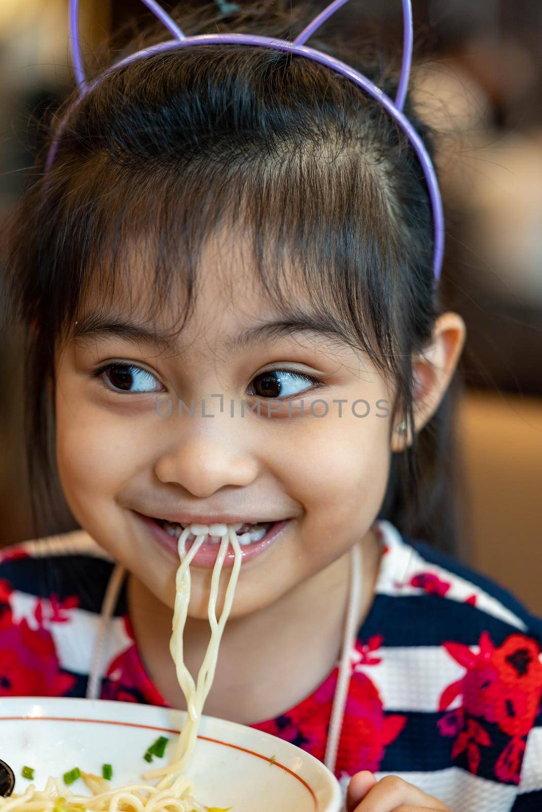 Female asian child while eating noodles. Child eating ramen noodles smiling and enjoying the food. Child eating spaghetti by billroque