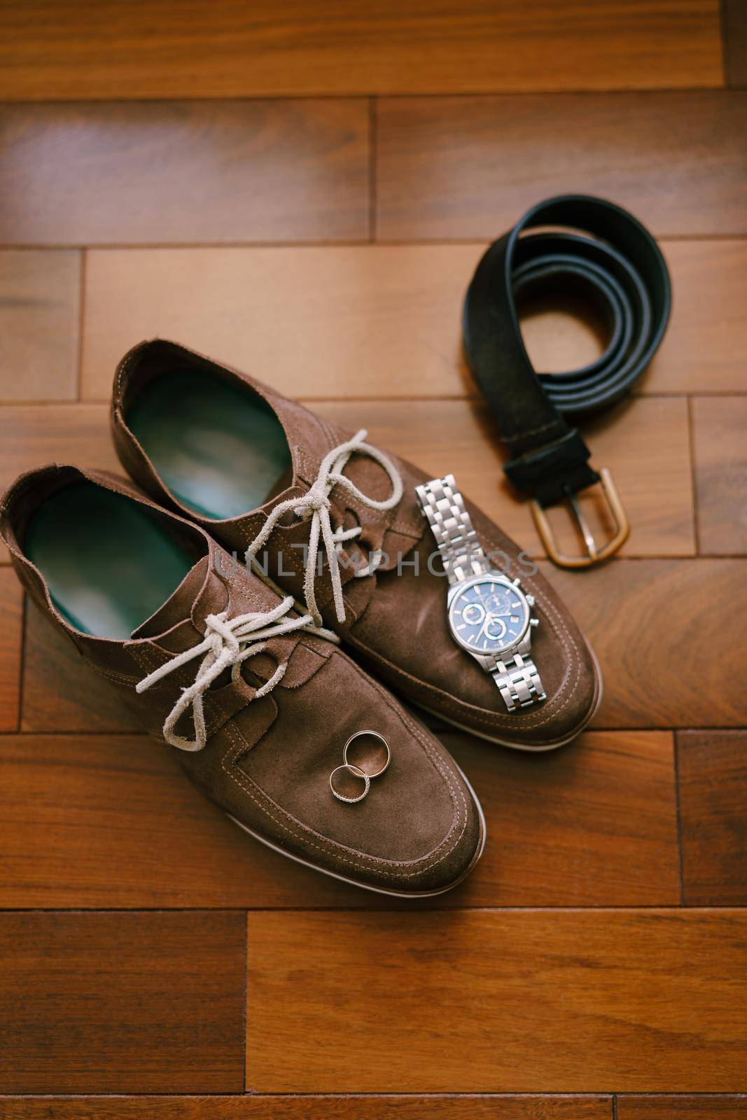 Brown mens suede shoes with wristwatch, belt and wedding rings of the bride and groom on a wooden texture. High quality photo