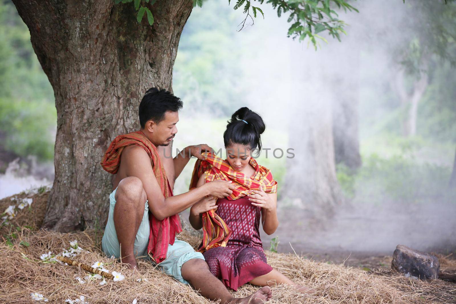 Couple love of Asian Young man and women sitting under tree against buffalo and natural background, rural way of life in the Northeast of Thailand. A young man was blowing a bamboo mouth organ to his lover. by chuanchai