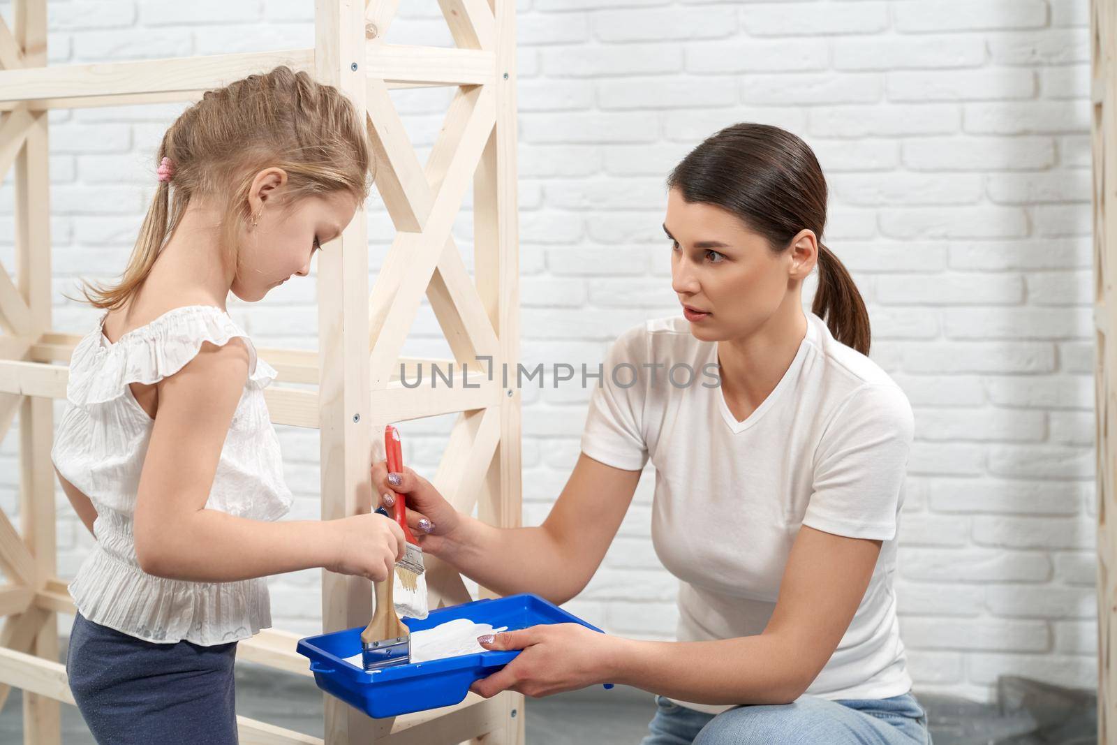 Close up of mother and daughter holding brushes and white paint. Concept of preparing painting rack.