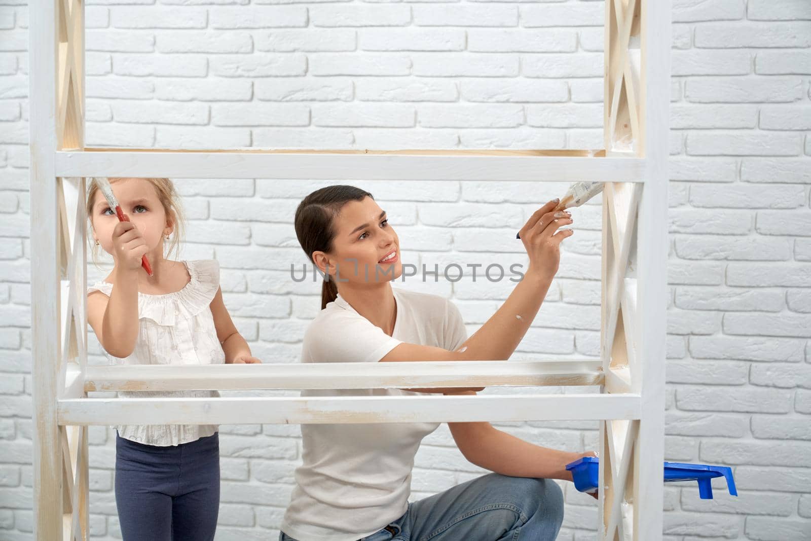 Close up of smiling mother with daughter painting rack in white color at home. Concept of repair at home.