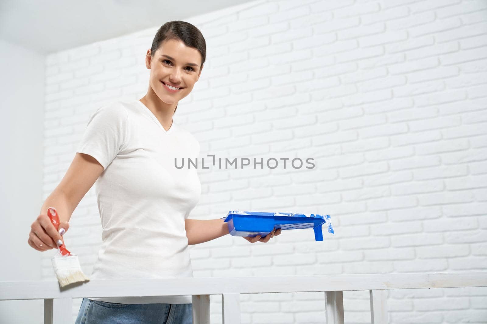 Smiling brunette woman painting rack in white color in empty room. Concept of repair at home.