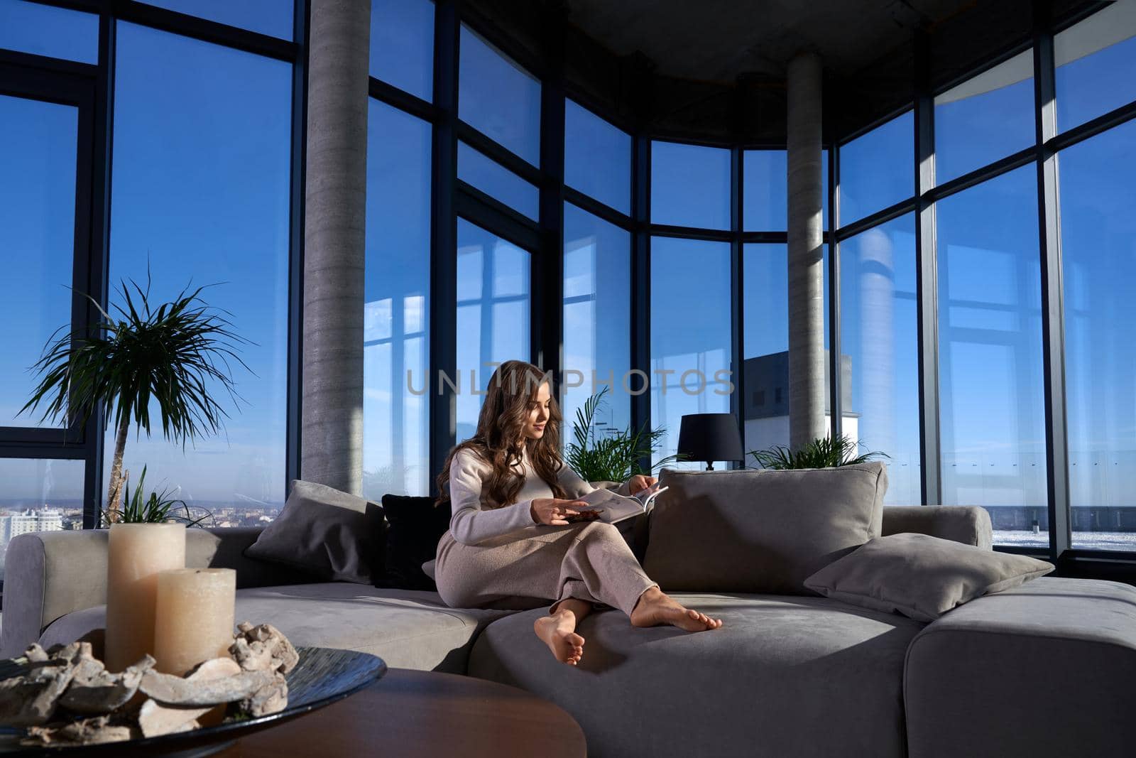 Front view of young brunette woman sitting on cozy grey sofa reading book near large panoramic window with incredible view on city. Concept of relaxing at home or hotel with modern interior.