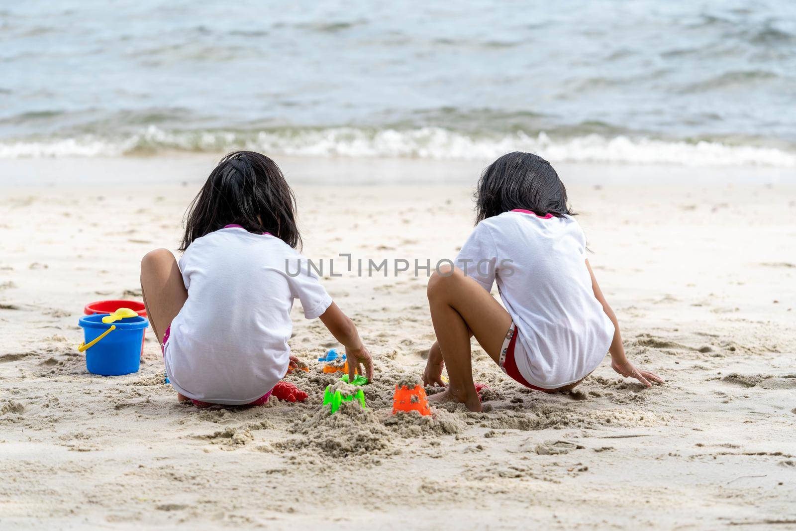 Twin girls while playing beach toys and sitting on a beach sand with white sand and waves by billroque