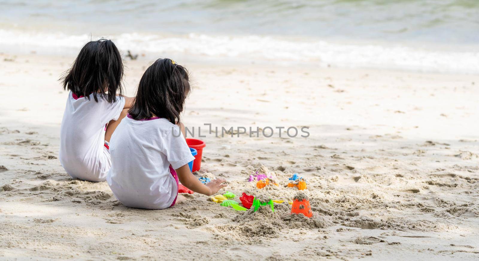 Twin girls while playing beach toys and sitting on a beach sand with white sand and waves