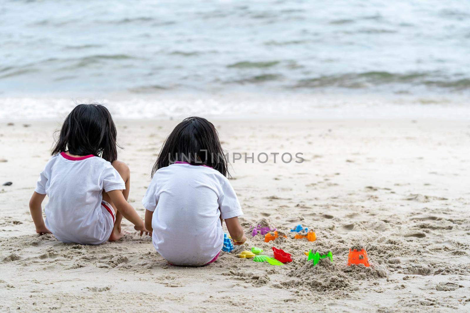 Twin girls while playing beach toys and sitting on a beach sand with white sand and waves by billroque