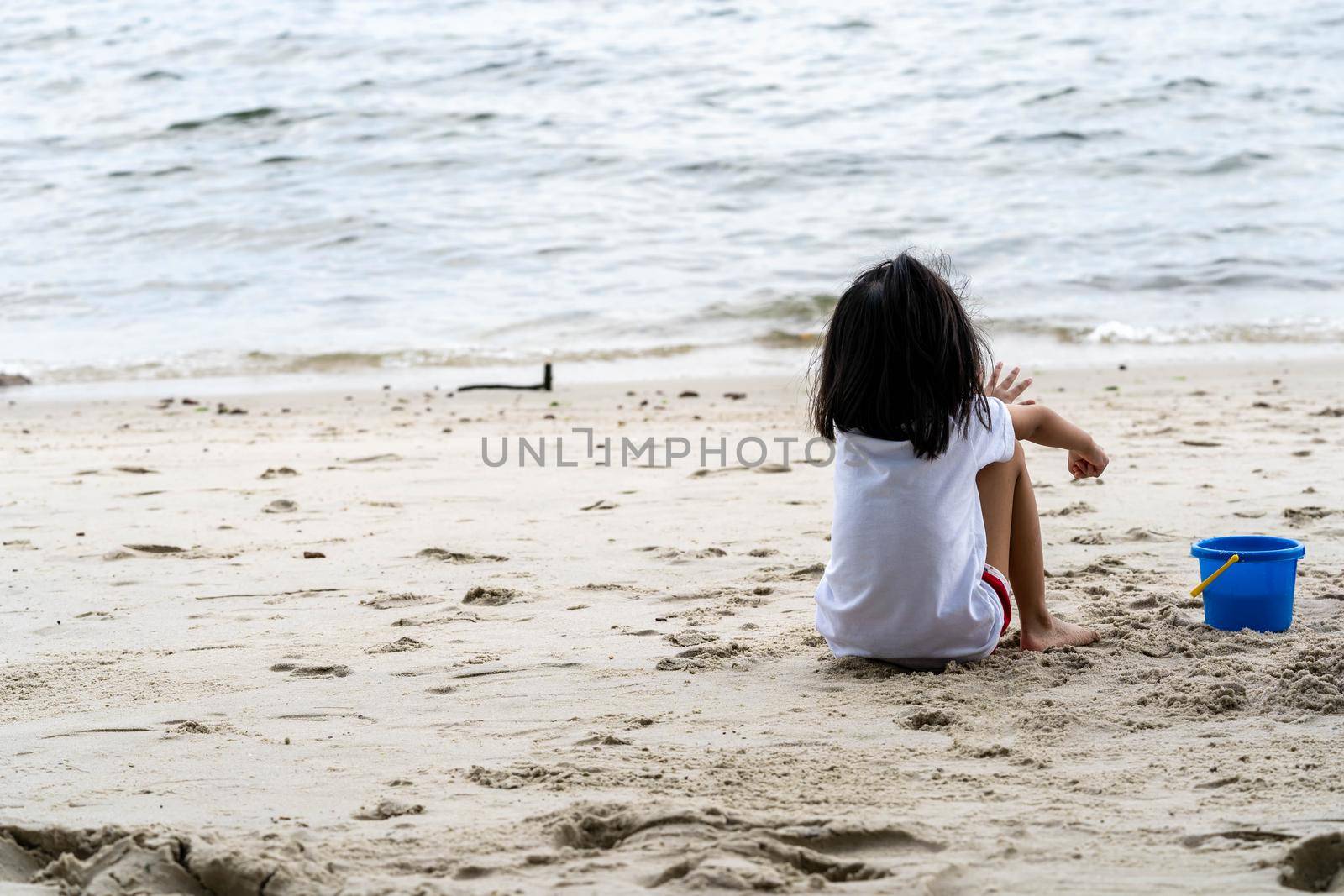 Little girl while sitting on a sand in a beach and looking at the ocean alone by billroque