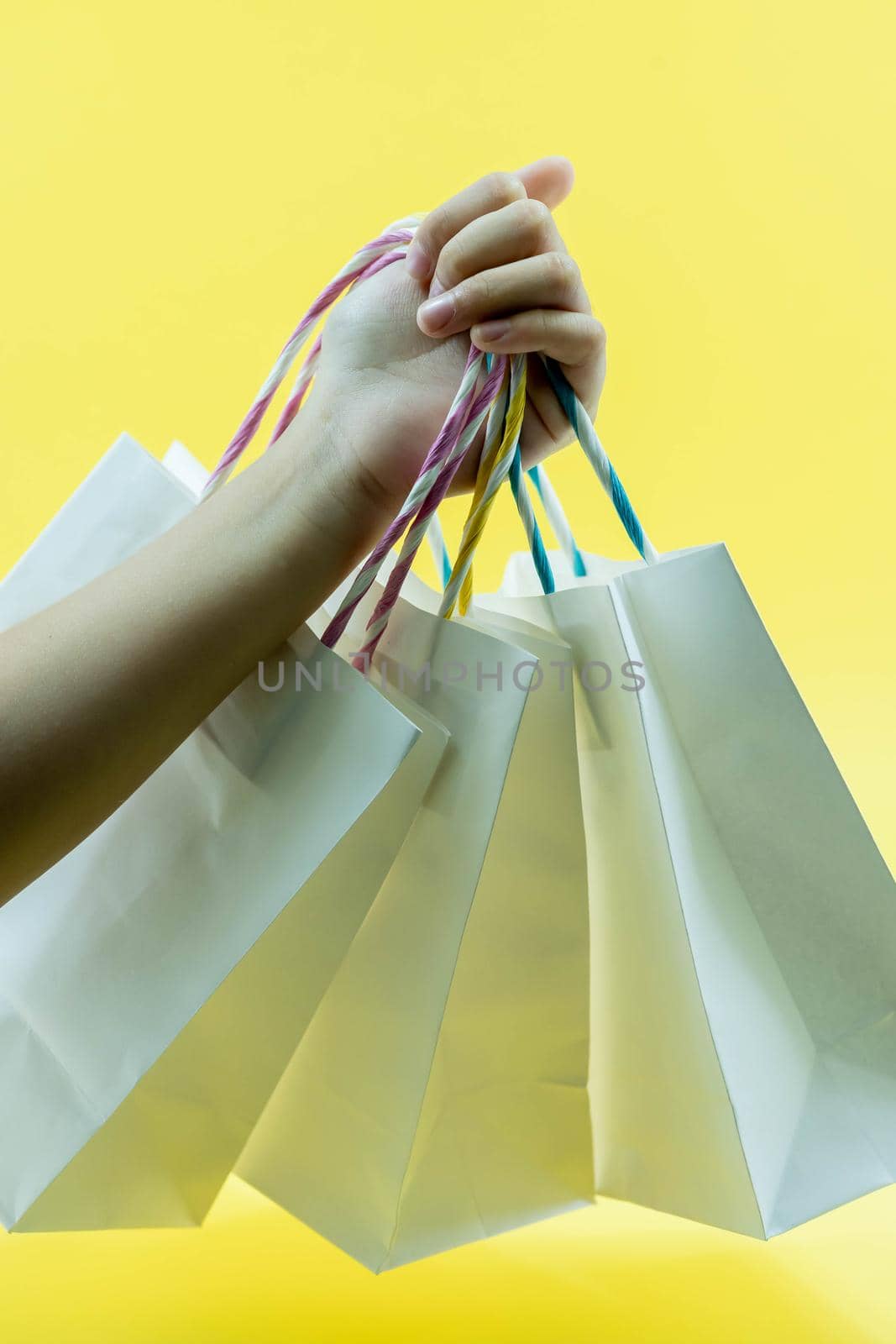 Cropped shot of child hand holding bunch of different colorful blank shopping bags over isolated Yellow background. Black friday sale concept. Copy space, close up.