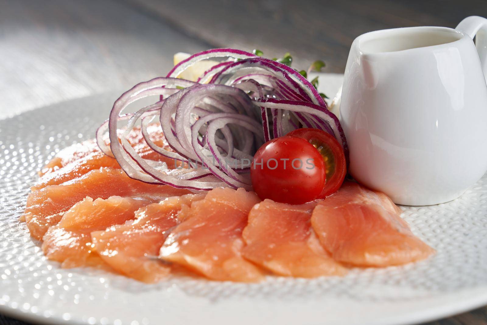 Close up view of delicious raw salmon slices on white plate served with lemon, onion, tomatoes, sprouts and small jar of oil. Tasty appetizer on wooden table in luxury restaurant for dinner.