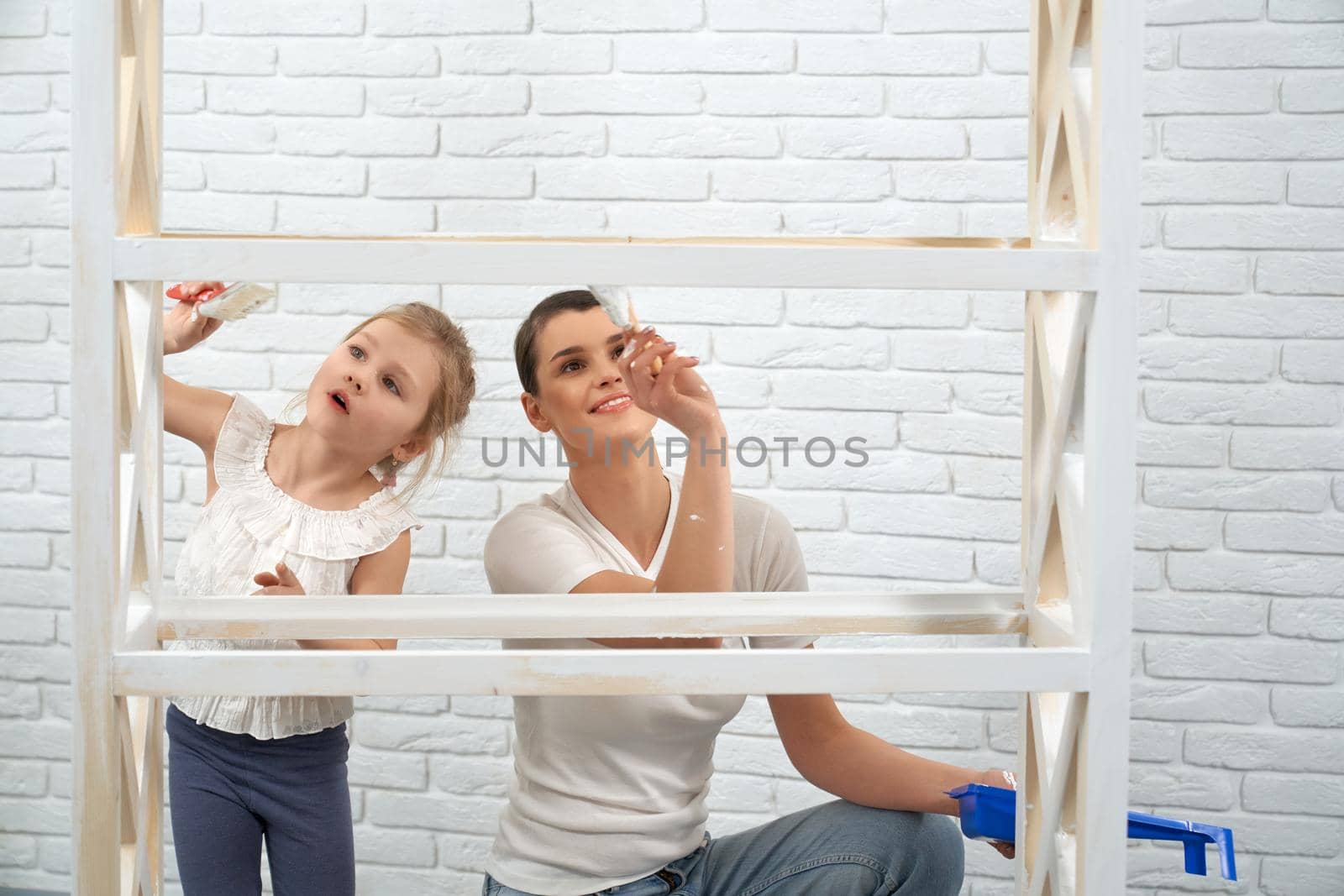 Close up of mother with little daughter painting wooden rack in white color. Concept of repair at home.