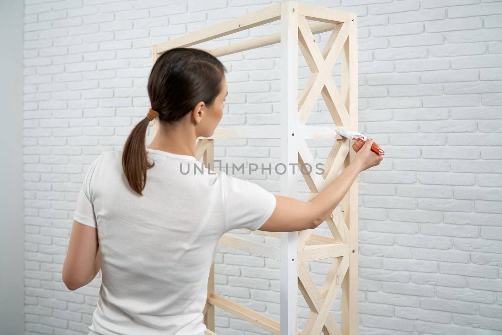 Close up of brunette woman holding brush and painting small wooden rack in white color. Concept of repair at home.