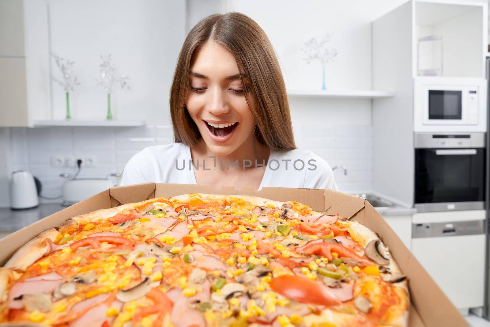 Happy young woman holding hot pizza in box at home. Concept of dinner in kitchen.