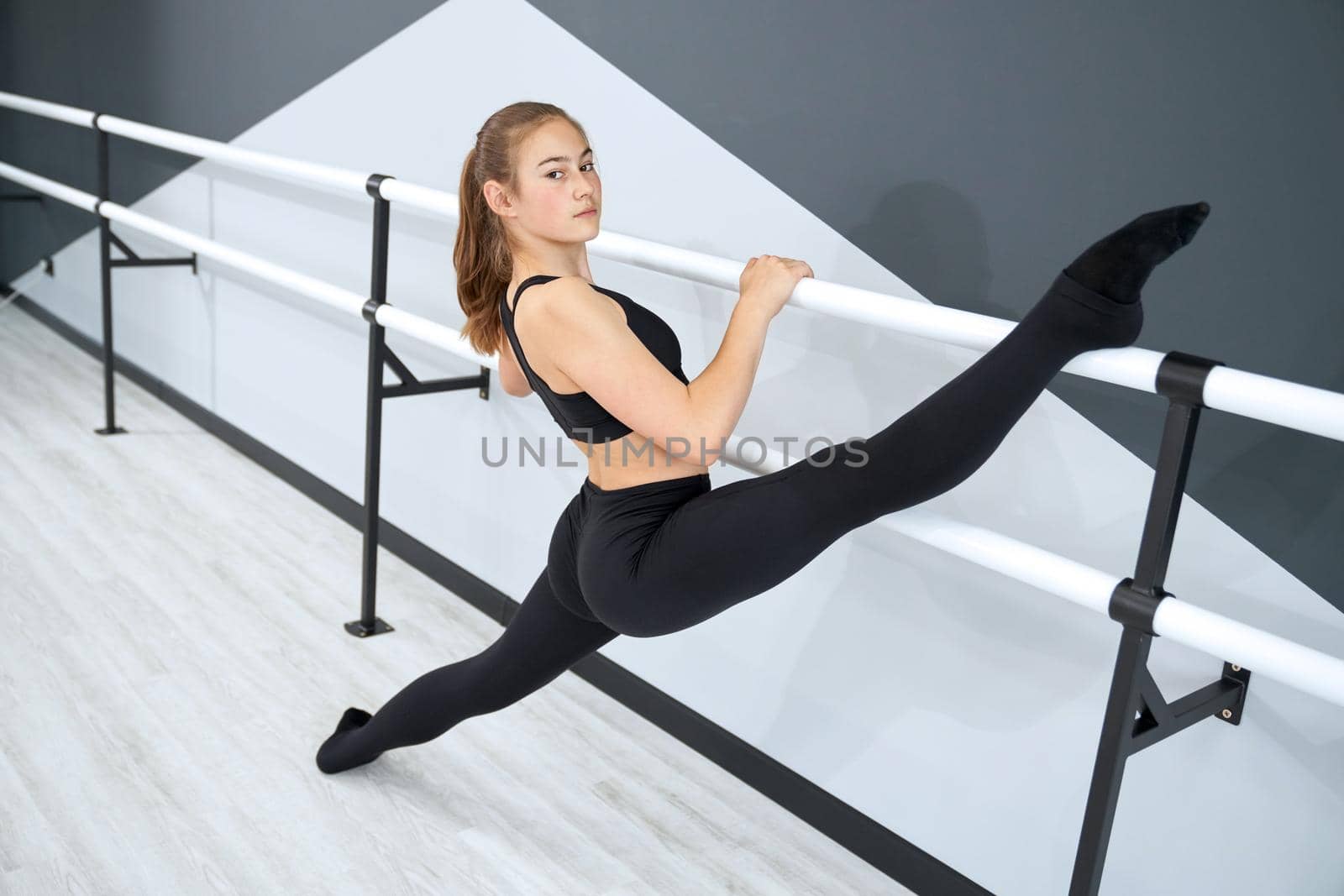 Beautiful teenager holding leg on handrail, looking at camera. Flexible female athlete wearing tight black sports clothes practicing split in empty dance hall. Concept of gymnastics, sport.