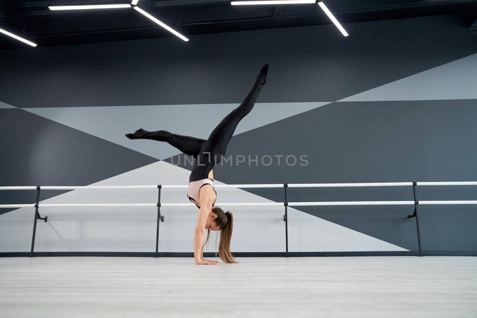 Strong fit woman in tight sportswear standing on arms and practicing split in air. Side view of flexible female dancer training before competition in big empty dance hall with ballet handrails.