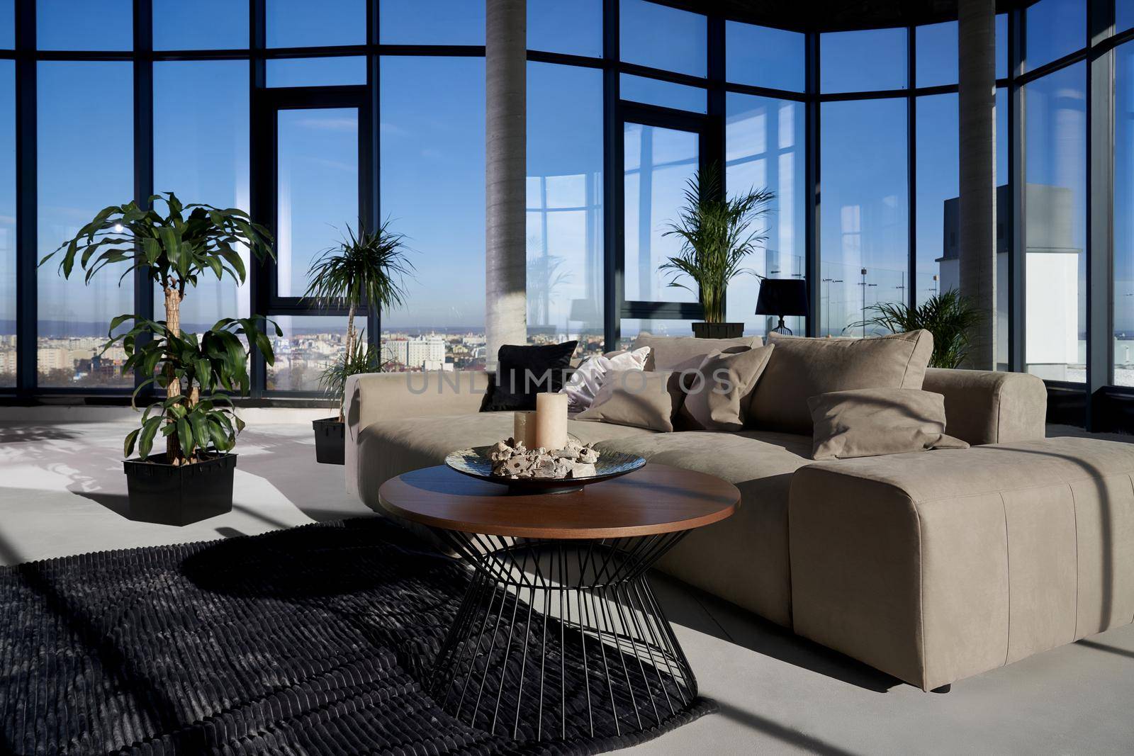 Front view of fashionable interior spacious room with large panoramic window with incredible view on city and cozy grey sofa with pillows and green flowerpots. Concept of modern interior.