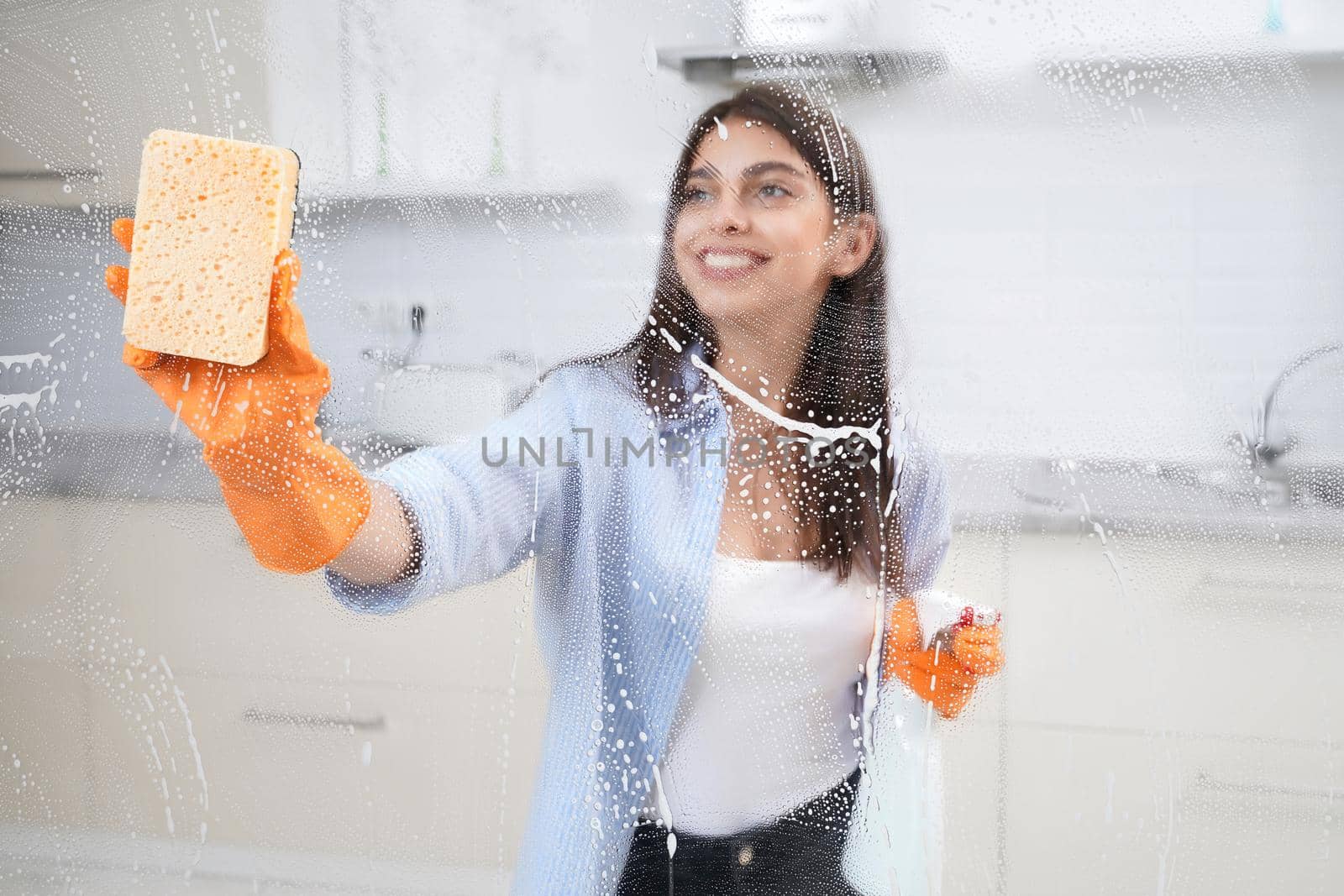 Smiling brunette woman washing window in rubber gloves with rag and detergent. Concept of cleaning window or glass.