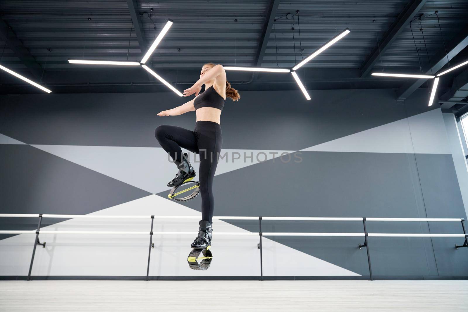 From below view of active female teenager doing cardio workout in hall, hi tech interior. Young pretty girl wearing black sportswear practicing dance moves while doing kangoo jumps and smiling.