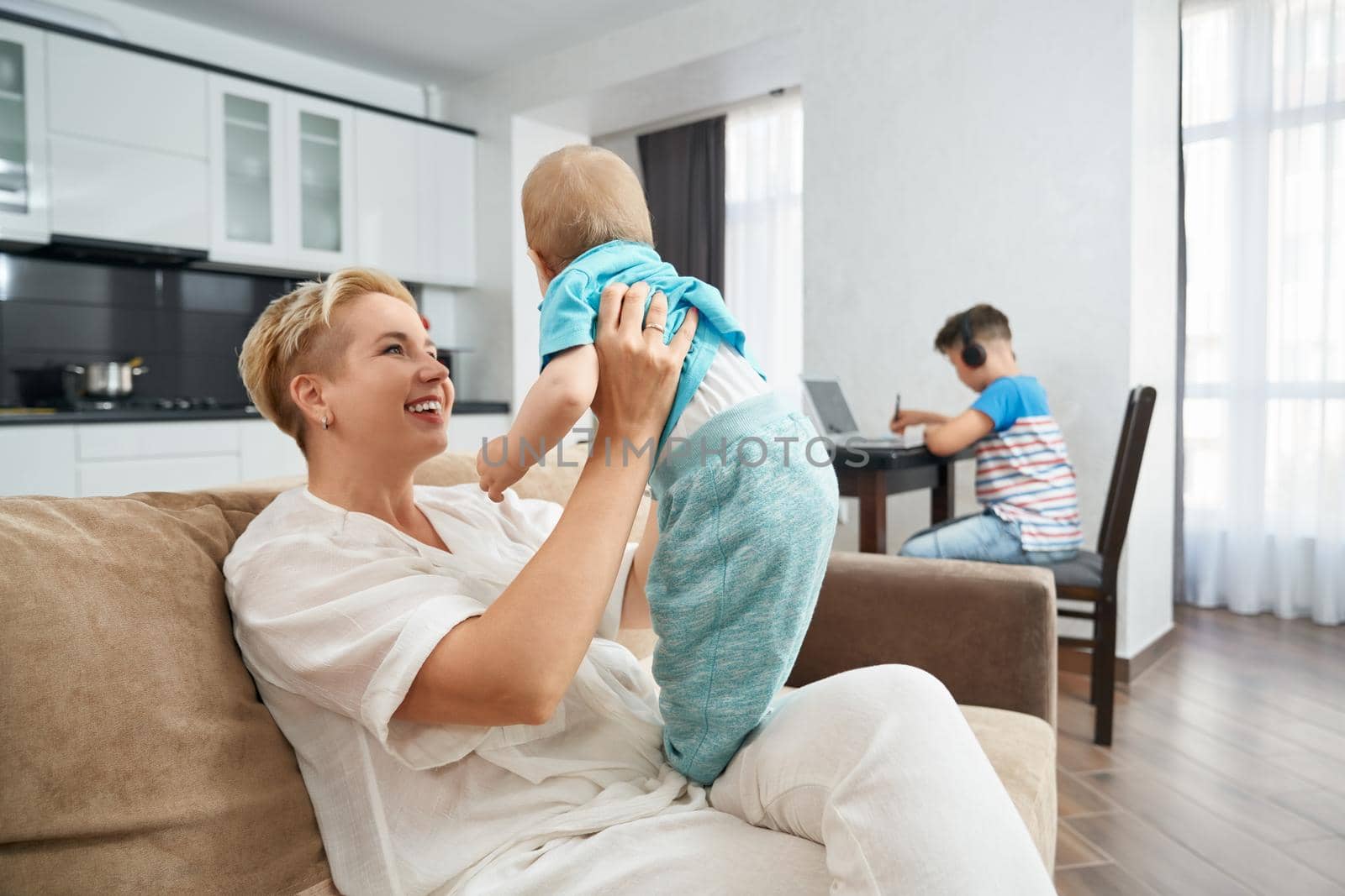 Smiling woman with short blond hair taking care of her little son while eldest son sitting at kitchen table and studying on laptop. Concept of family and technology.