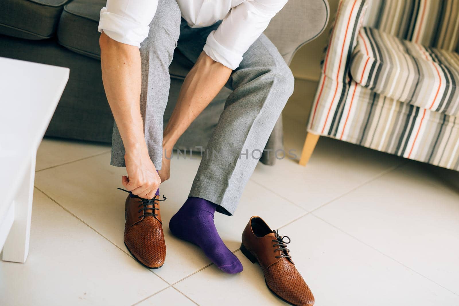 The man in gray slacks and a purple dress socks brown shoes with laces sitting on the couch.