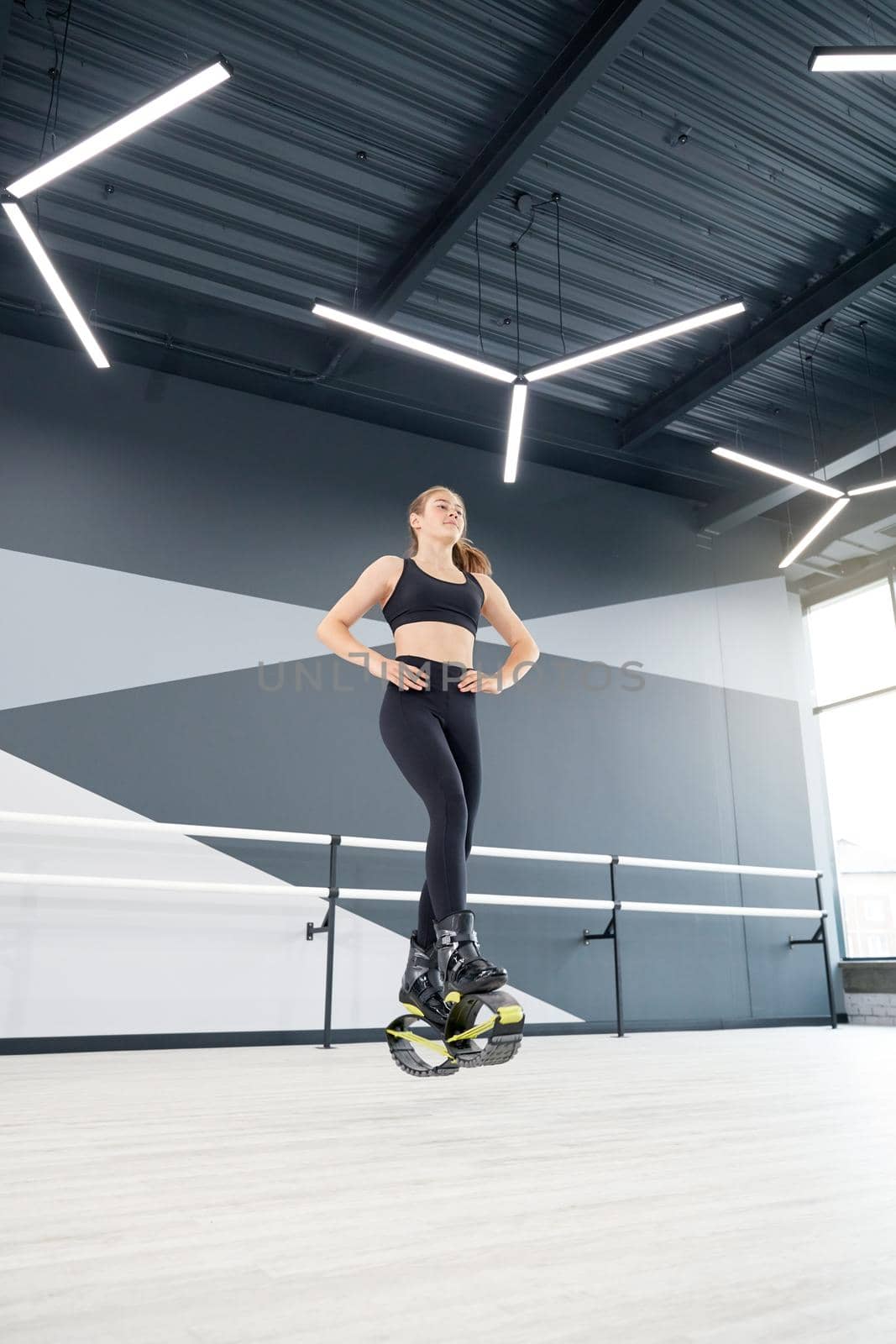 From below view of girl practicing dance moves while doing kangoo jumps, hands on waist. Young female athlete with ponytail wearing tight black sportswear, cardio exercise in hall, hi tech interior.
