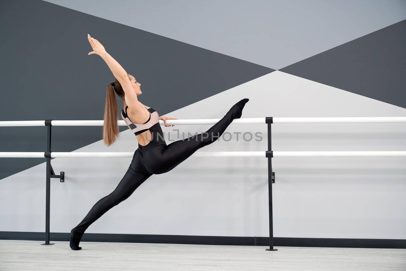 Fit flexible female athlete practicing in dance studio, hi tech interior. Side view of gorgeous adult woman with ponytail stretching leg on handrail, doing split. Concept of gymnastics.