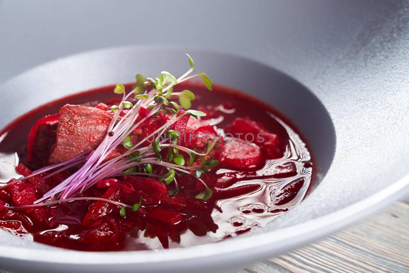 Close up view of bright red borsch, beet soup with meat and vegetables served with young green sprouts. Tasty hot dish in white plate in restaurant. Concept of traditional ukrainian cuisine.