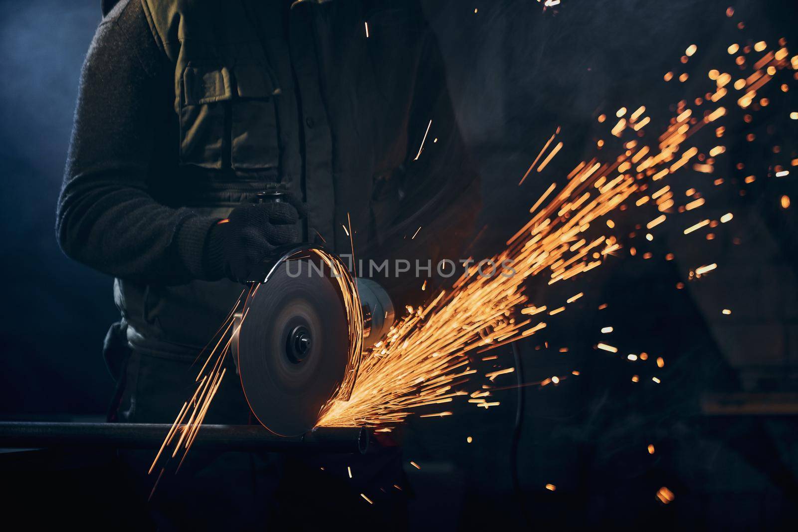Close up of man in special dark suit and black protective gloves working angle grinder for metal with large flash sparks. Concept of process working electric steel cutter machine.