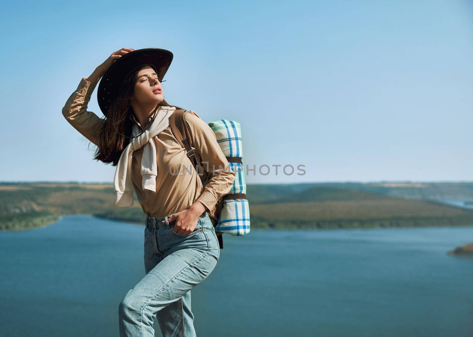 Pleasant young woman in casual outfit and cowboy hat enjoying sunny weather during hiking at Bakota area. Beautiful nature around near Dniester river.