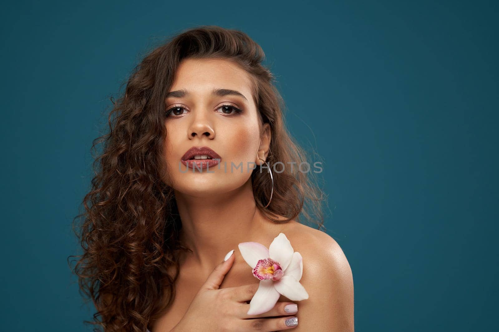 Headshot of young beautiful brunette female model holding orchid on naked shoulder, looking at camera. Pretty caucasian woman posing holding pink flower in hand. Beauty, youth concept.