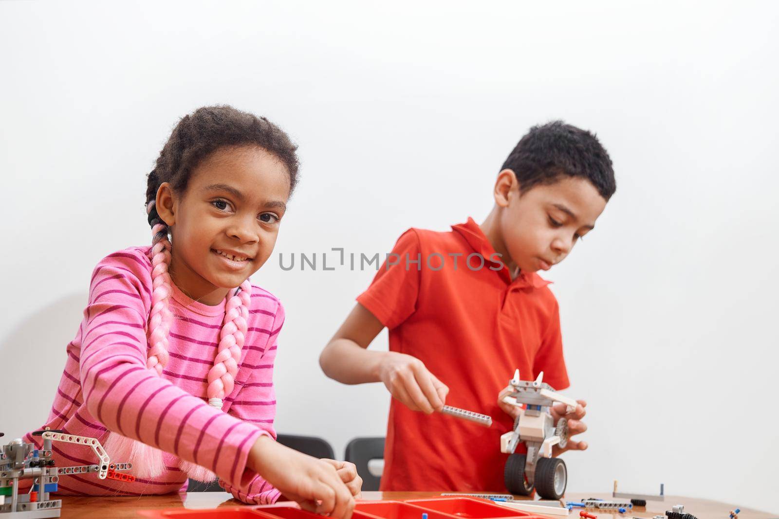 Front view of building kit for group of kids creating toys, having positive emotions and joy, selective focus of girl smiling and looking at camera. Close up of african friends working on project.