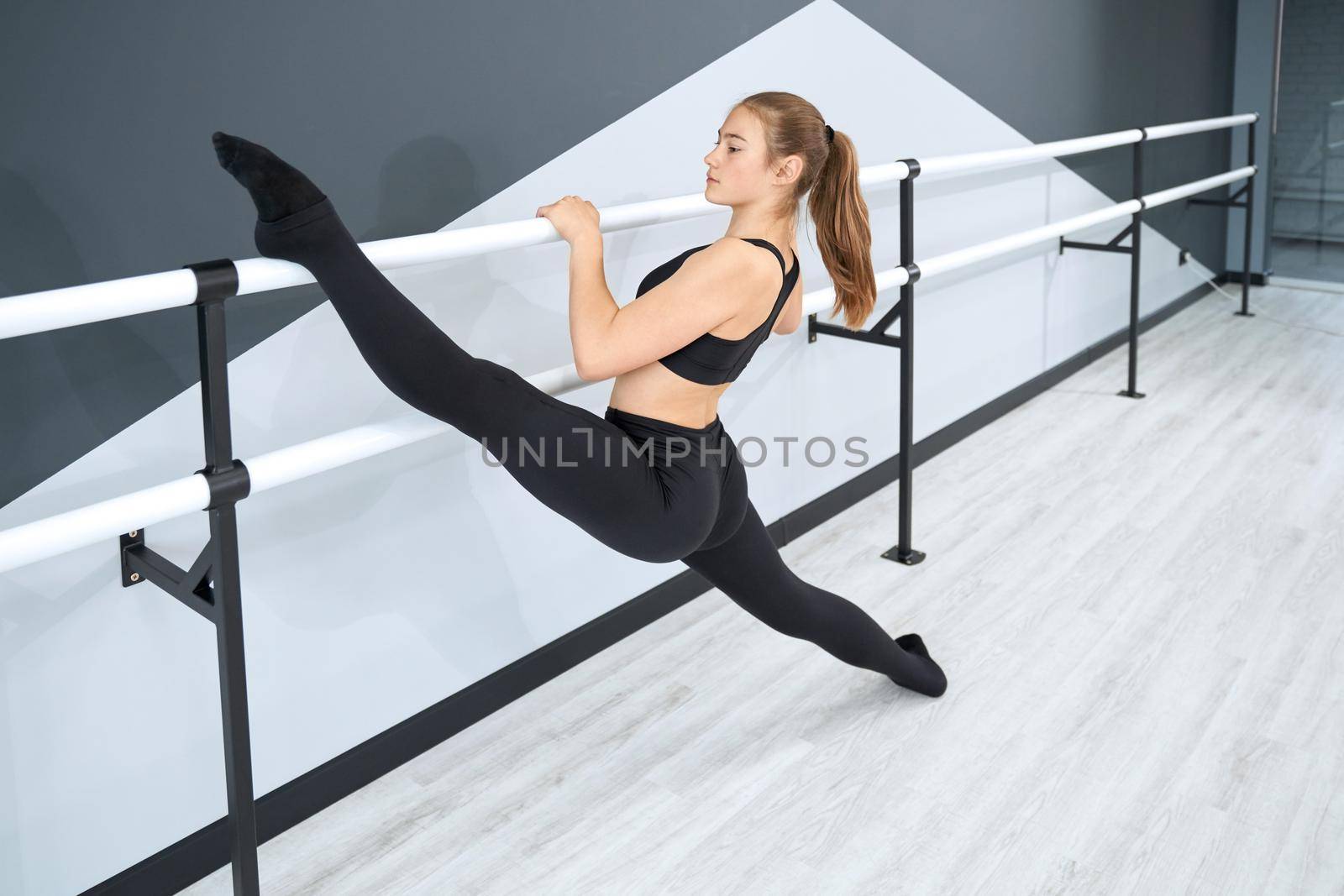 Side view of attractive teenager holding leg on handrail. Flexible female athlete wearing tight black sports clothes practicing split in empty dance hall with hi tech interior. Concept of gymnastics.