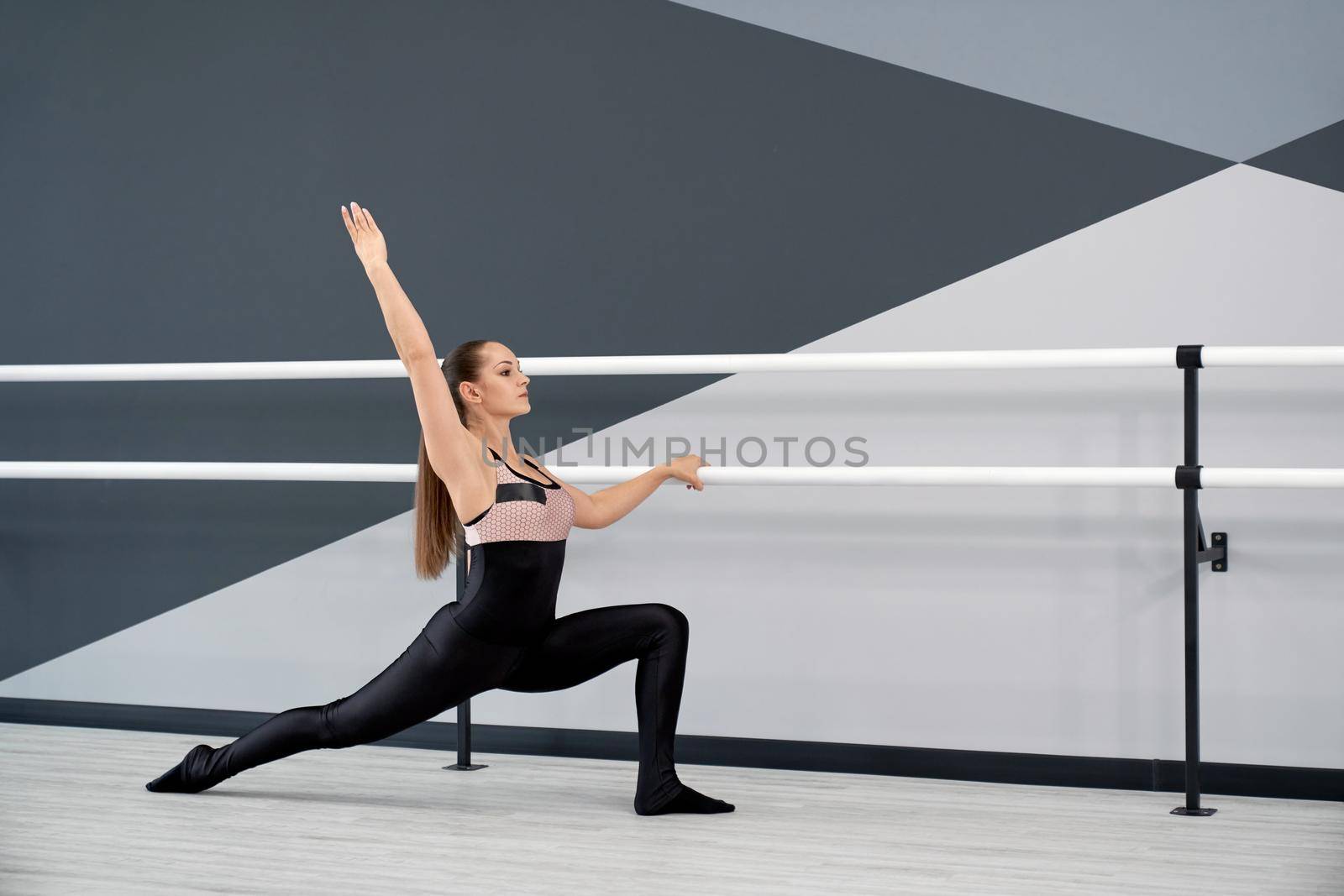 Side view of gorgeous flexible female athlete stretching with hand up in hall. Adult woman in tight outfit practicing lunges, holding handrails in ballet studio. Concept of choreography, gymnastics.