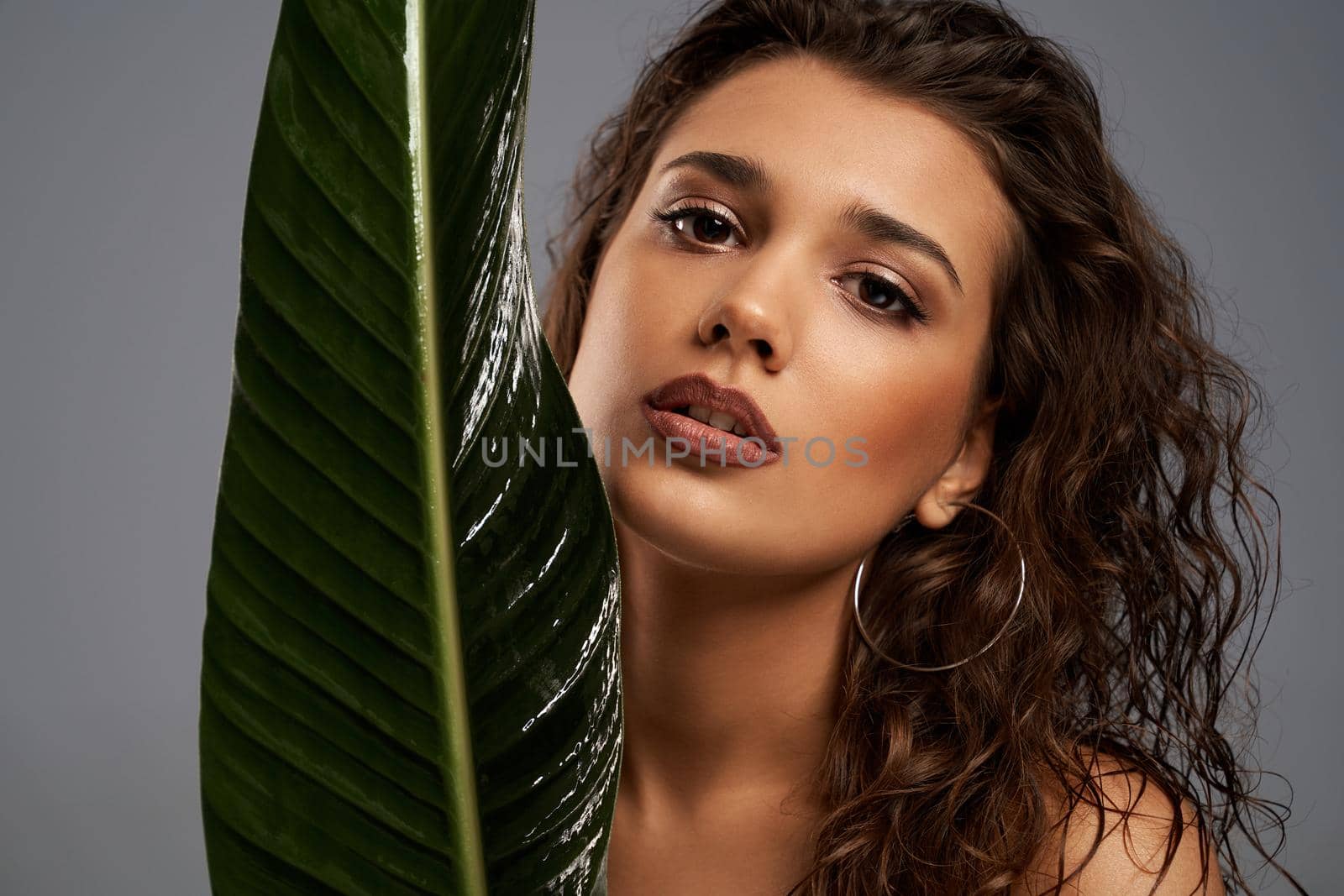 Close up of beautiful naked female model looking at camera. Crop portrait of young brunette woman with perfect makeup posing with mouth open, holding big green leaf, isolated on gray.