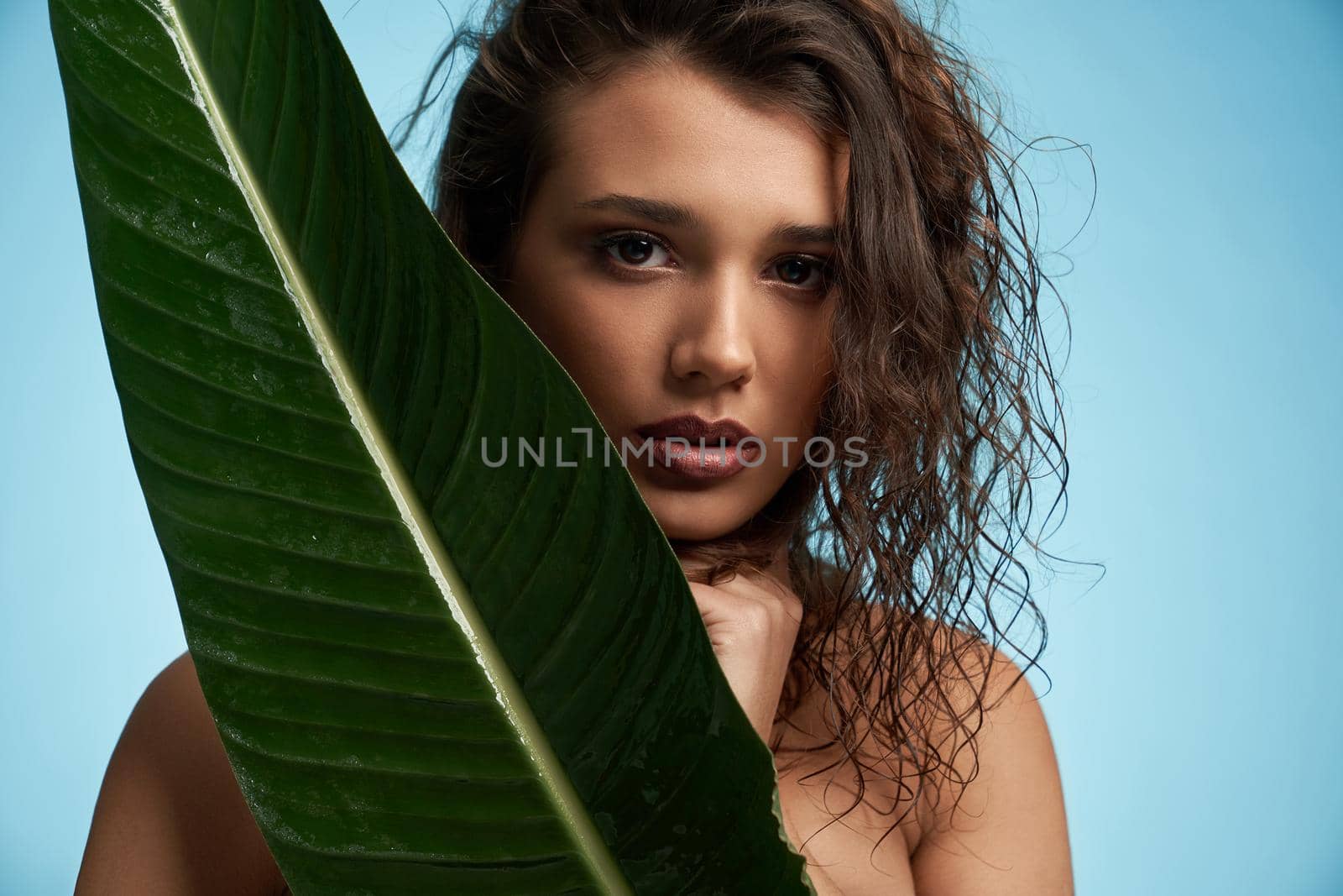 Close up portrait of astinishing naked female model looking at camera. Crop of young brunette woman with perfect makeup and fresh skin posing with mouth open, holding big green leaf. Beauty concept.