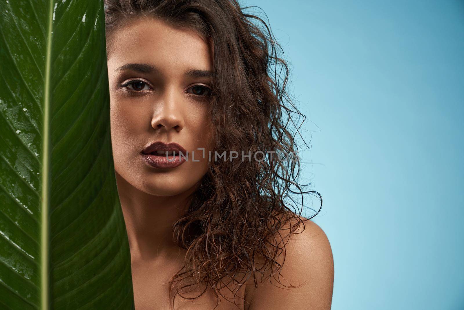 Close up of beautiful naked female model looking at camera. Crop portrait of young brunette woman with perfect makeup posing with mouth open, holding big green leaf, isolated on blue.