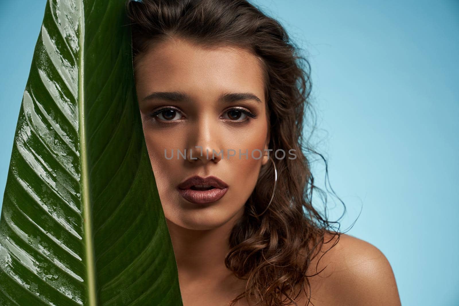 Close up portrait of astinishing naked female model looking at camera. Crop of young brunette woman with perfect makeup and fresh skin posing with mouth open, holding big green leaf. Beauty concept.