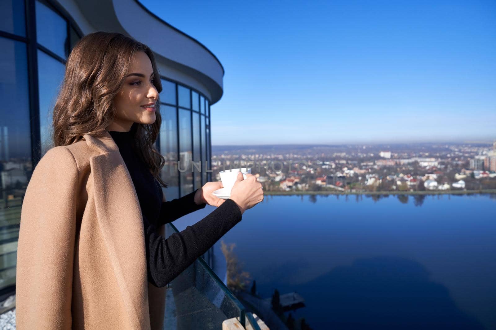 Gorgeous dark-haired woman enjoying beautiful view from high balcony of modern apartment. Beautiful lady enjoying morning time with cup of coffee.