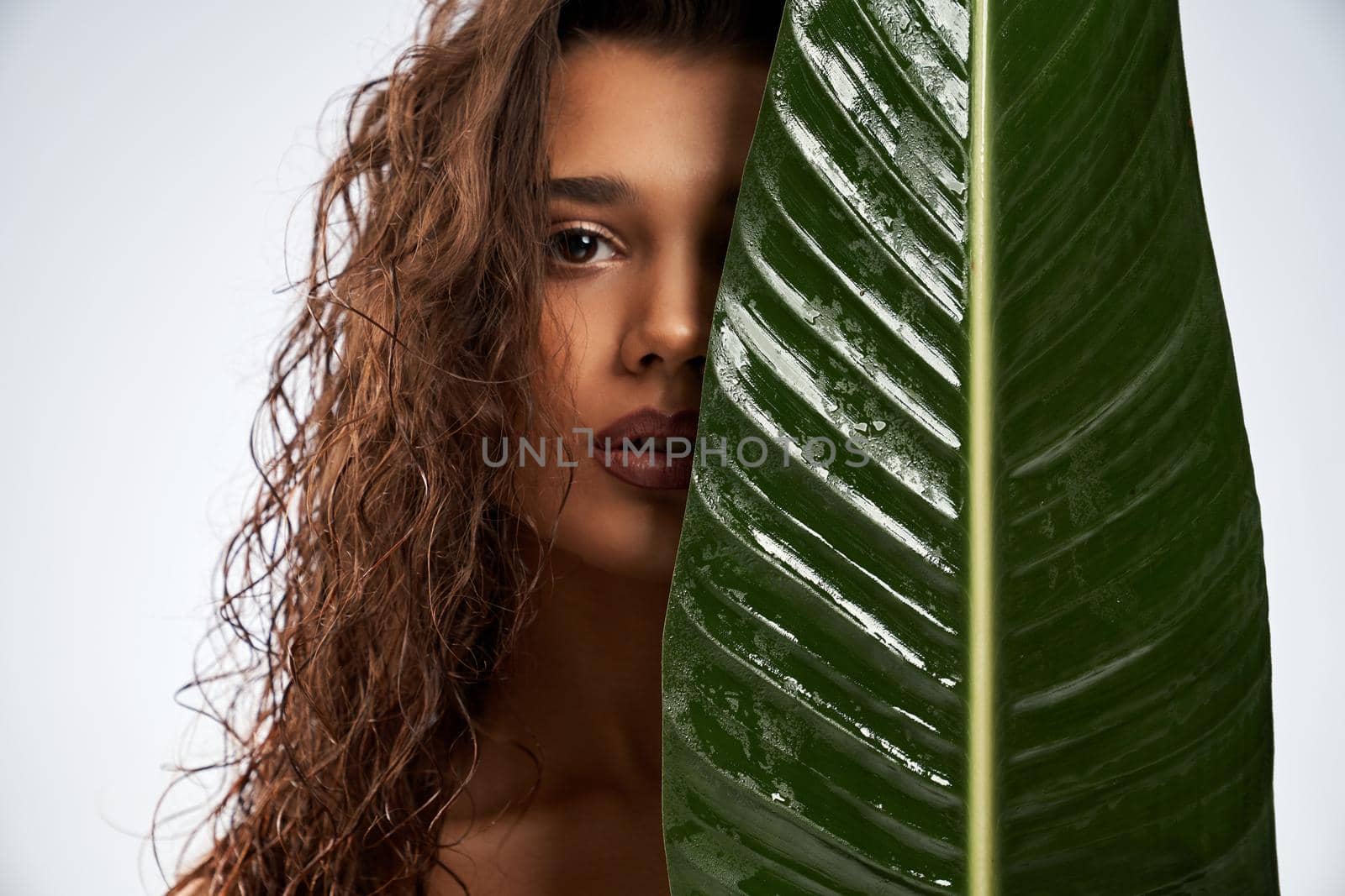 Close up of beautiful female model standing and looking at camera isolated on white. Portrait of young naked woman with perfect makeup posing with mouth open and hiding half of face behind green leaf.