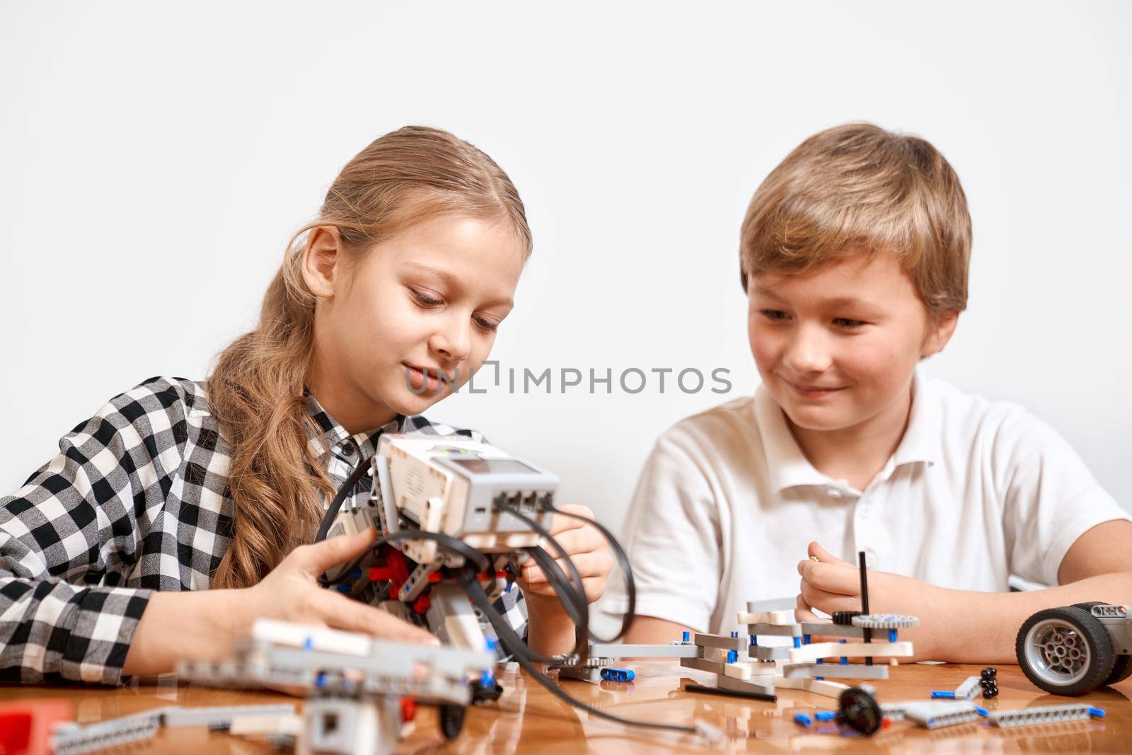 Front view of boy and girl having fun, creating robot. Science engineering. Nice interested friends smiling, chatting and working on project together using interesting building kit for kids on table.