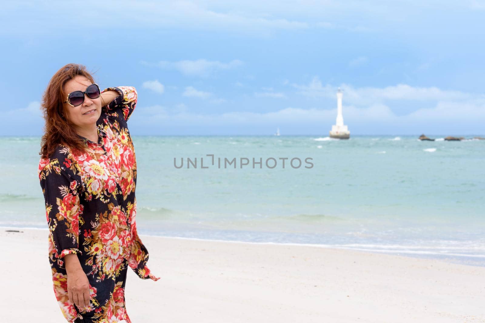 Middle-aged woman tourist wearing sunglasse looking at the camera and smiling on the beach and sea in summer sky background at Koh Tarutao island, Satun, Thailand