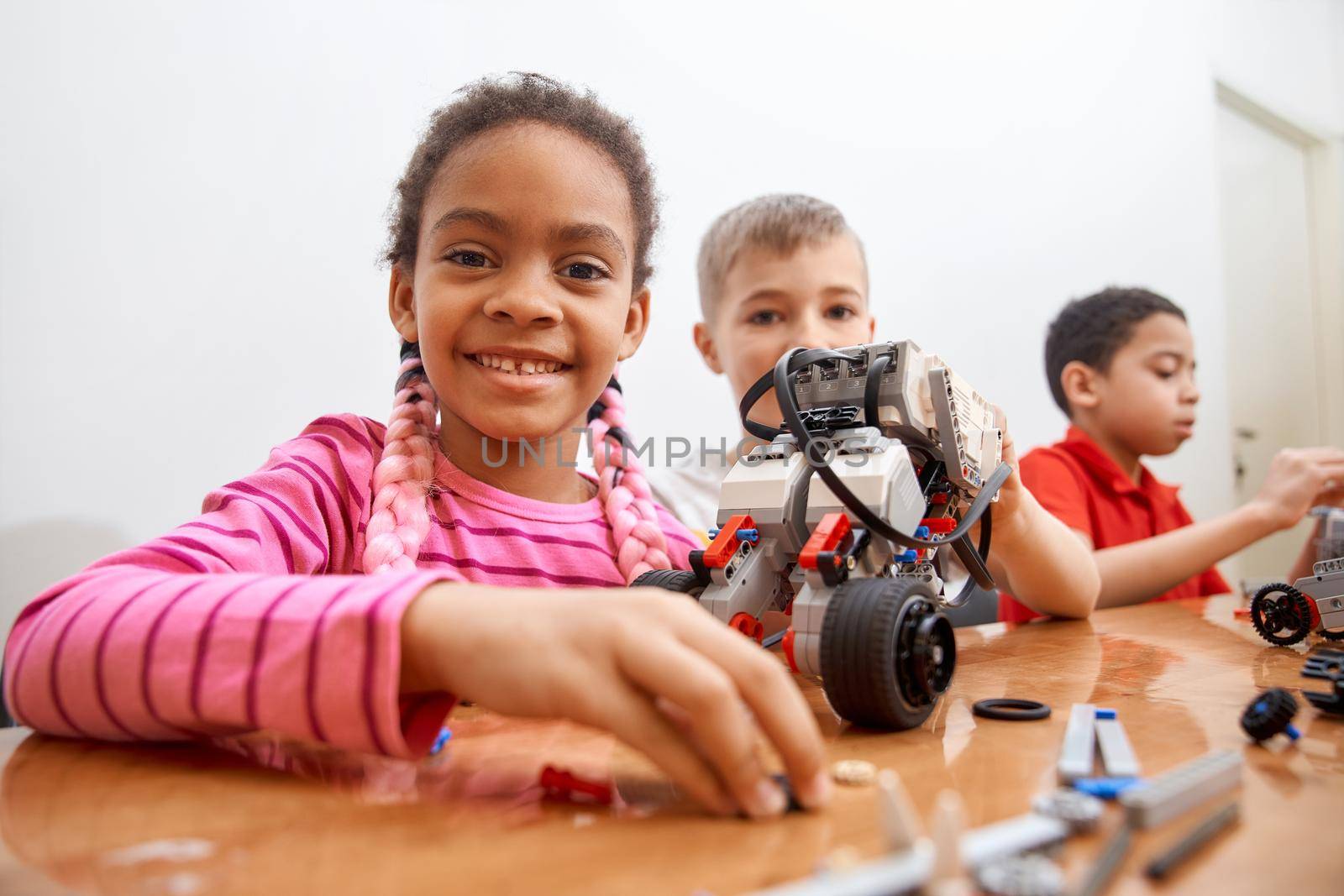 Front view of building kit for group of three multiracial kids creating toys, having positive emotions and joy, african girl smiling and looking at camera. Close up of friends working on project.